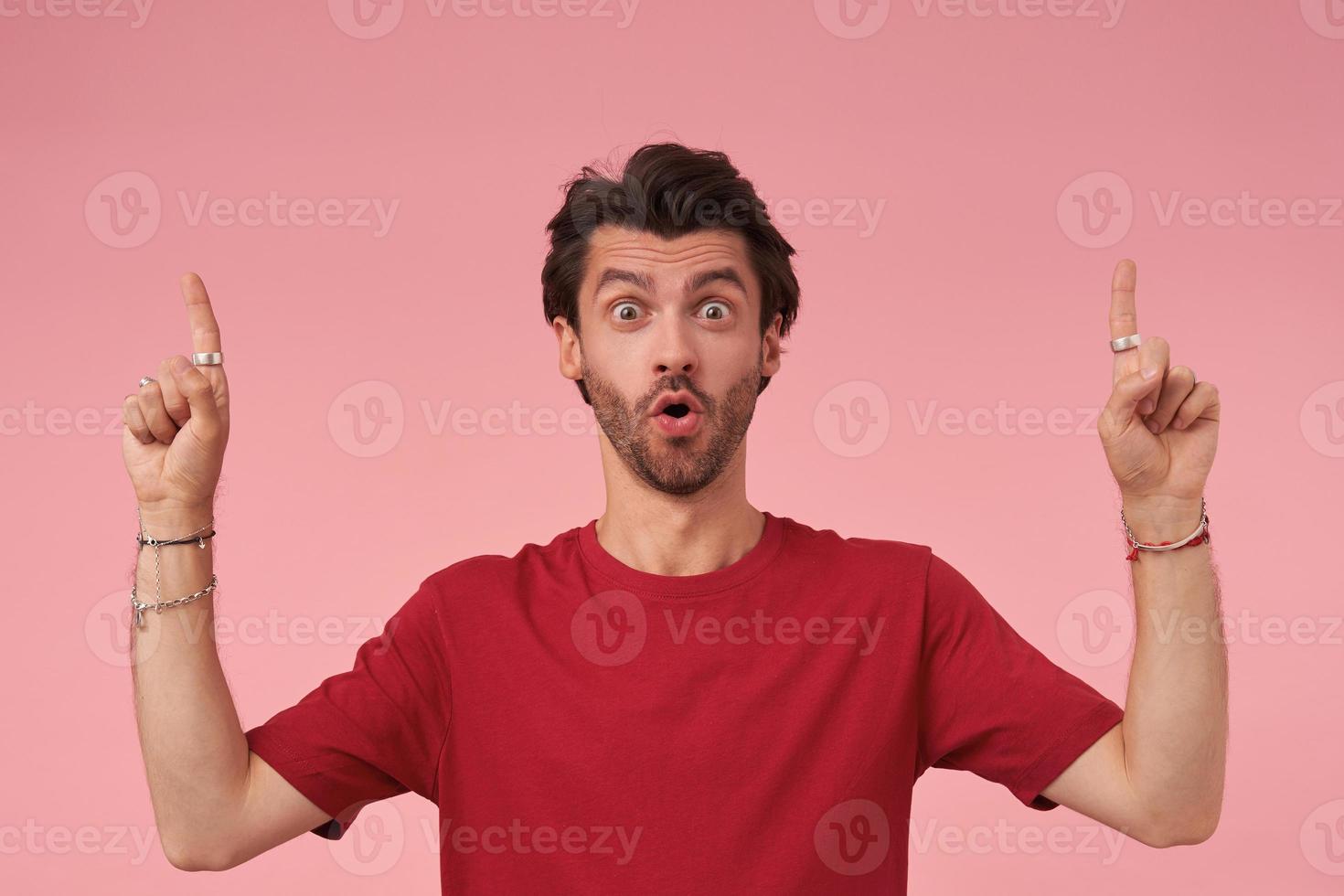 Amazed young man with beard in casual clothes standing over pink background with raised index fingers, looking at camera with wide mouth opened, contracting forehead and raising eyebrows photo