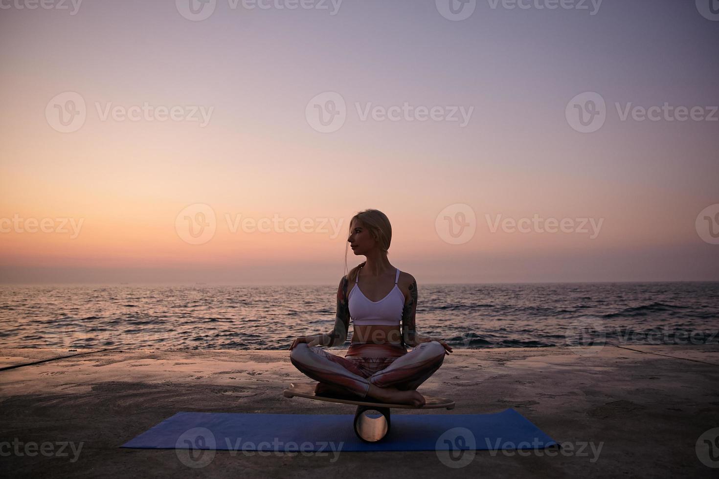 Outdoor photo of sporty blonde woman with casual hairstyle in sporty wear posing over sea view during sunrise, sitting on balance board with crossed legs and looking aside