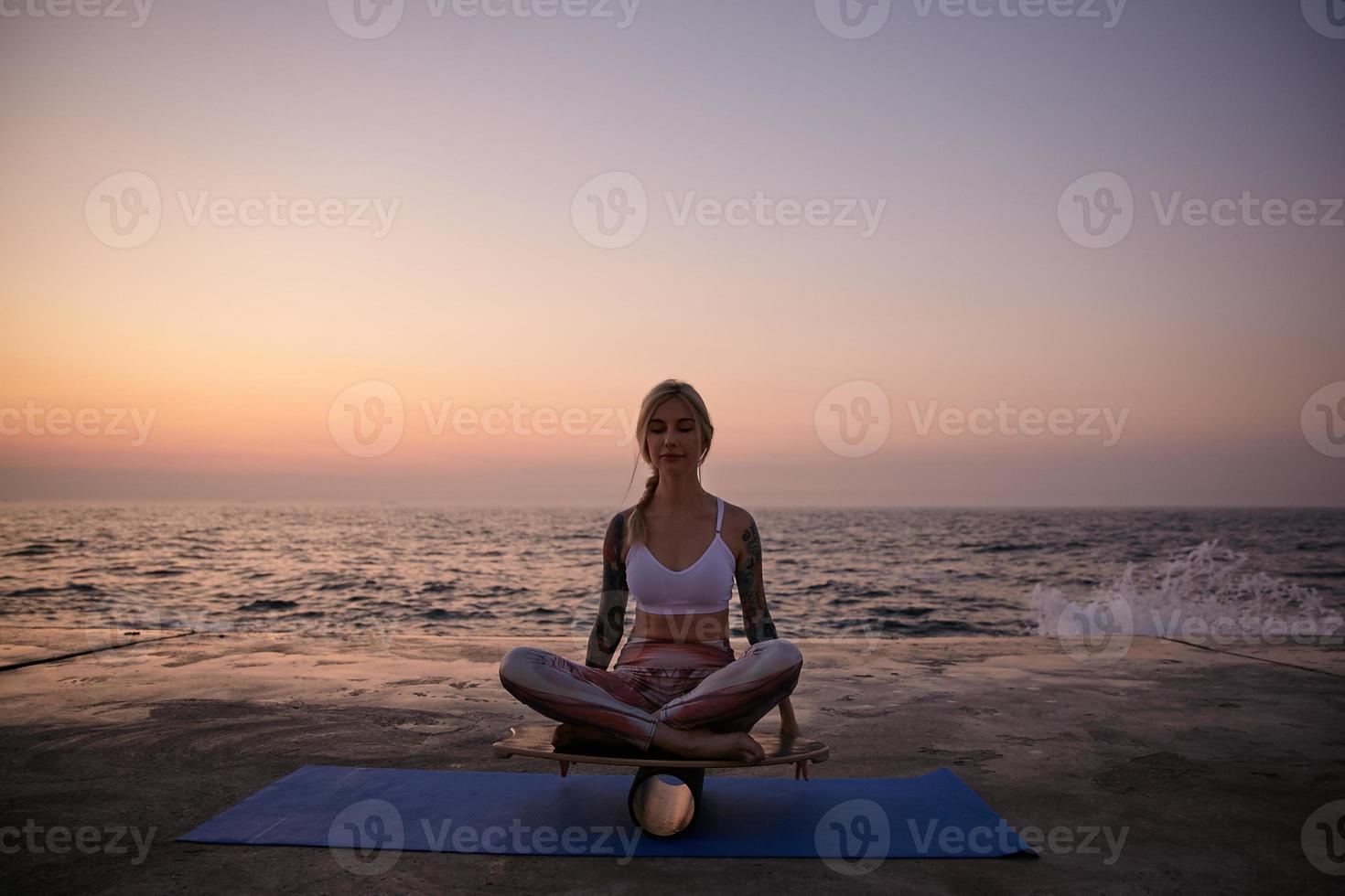 Outdoor photo of slim blonde woman sitting on balance board with closed eyes and calm face, posing over sea view during sunrise, helping to keep balance with hands
