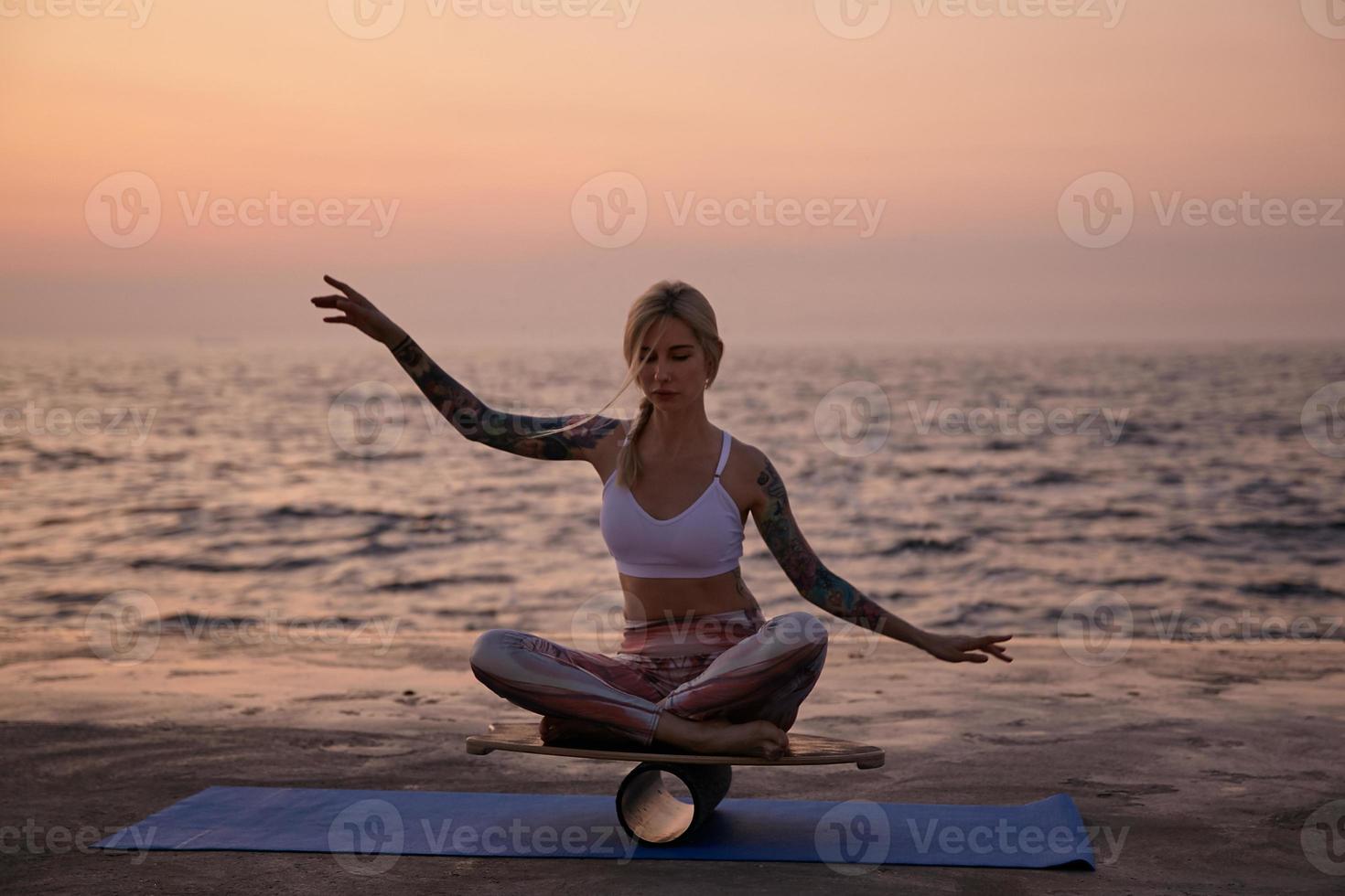 Young healthy woman with casual hairstyle sitting on wooden desk and balancing with hands, looking concentrated and calm, posing over seasde view on early morning photo