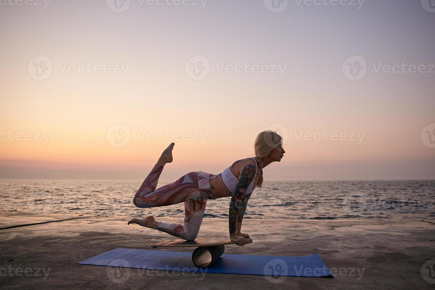 Outdoor shot of young blonde female with tattoos posing over sea view, having good body shape, wearing sporty top and leggins, making exercises with balancer, standing on wooden board during sunrise photo