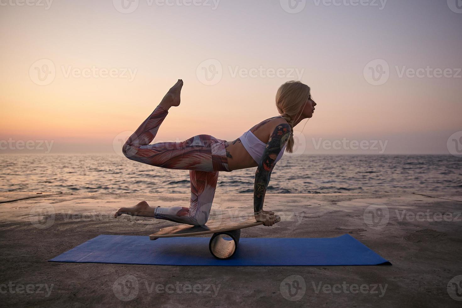 Young sporty blonde lady in good physical condition posing over sea view in sporty clothes, standing on special sports equipment with raised leg, balancing on wooden board outdoor on early morning photo