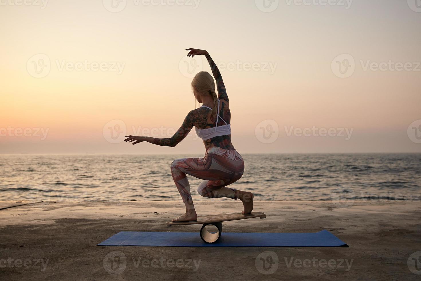 Outdoor shot of young tattooed female with good body shape posing over sea view, wearing sporty clothes, trying to keep balance on special sports equipment with raised hands photo