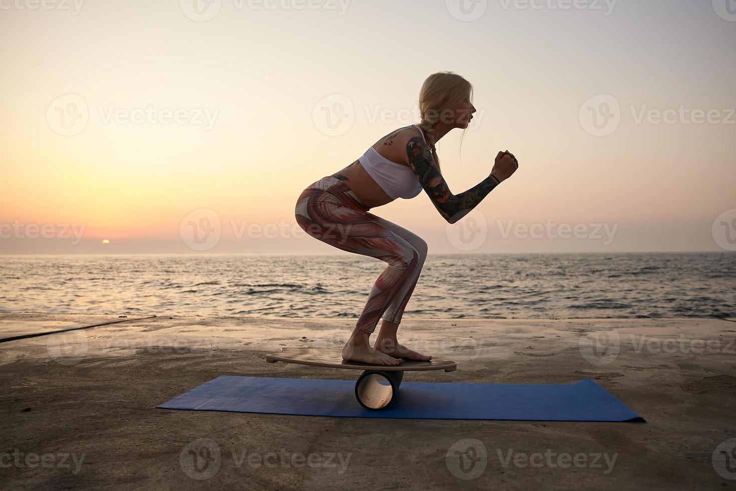 Outdoor photo of young sporty female with tattooes in sporstwear holding pose on balancer, posing over sea view during sunrise, standing on special sports equipment in half squat