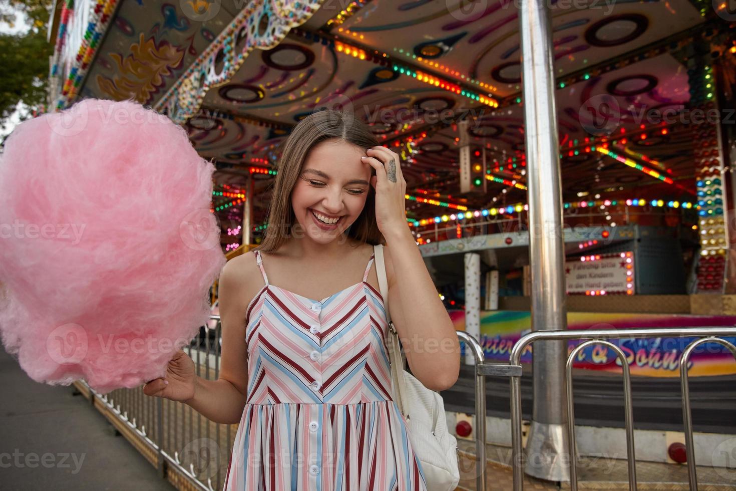 feliz joven dama atractiva con el pelo largo y castaño con un vestido claro con correas, de pie sobre un parque de atracciones con algodón de azúcar rosa en la mano, tocando la frente con los ojos cerrados y sonriendo ampliamente foto