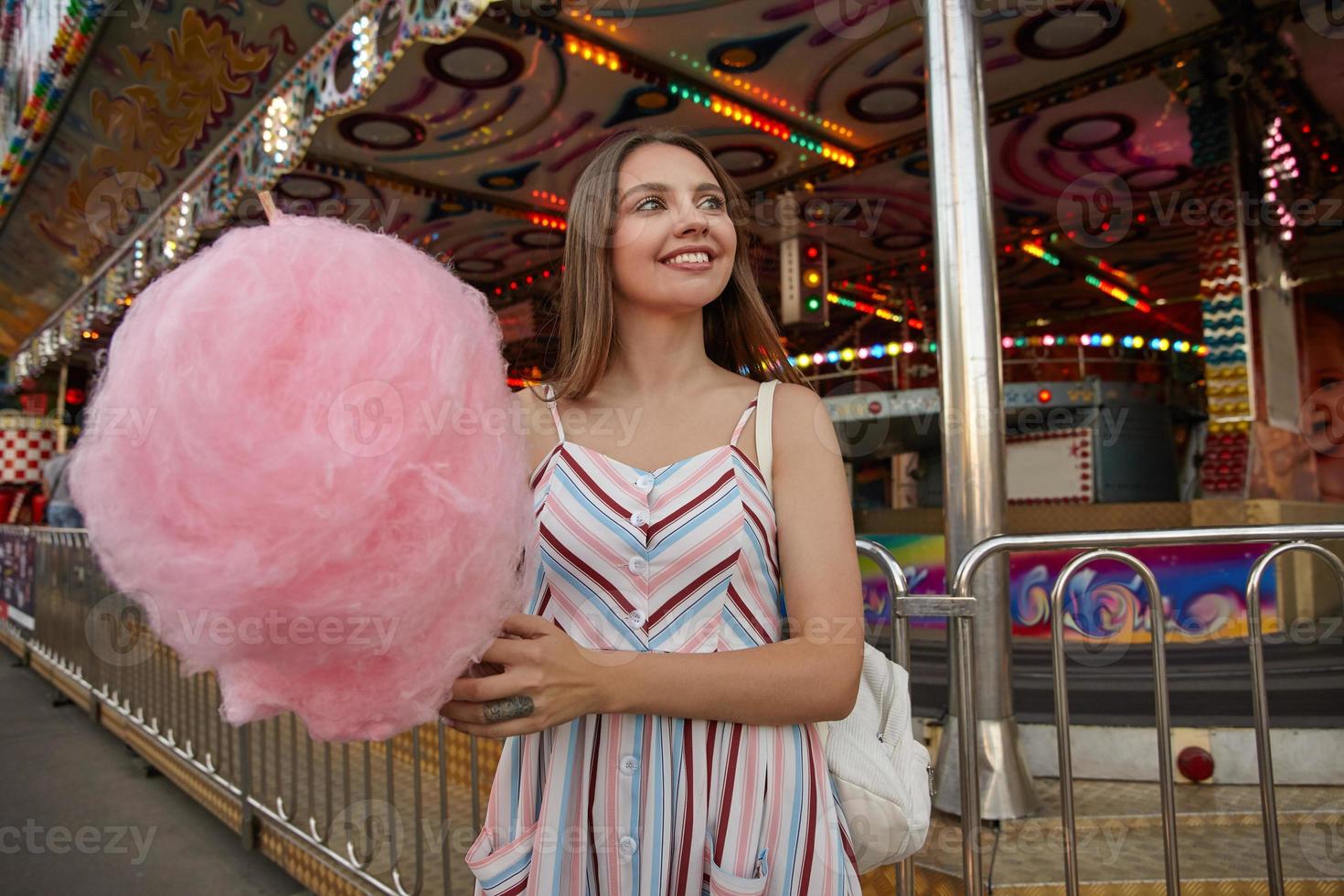 Outdoor photo of lovely young brunette female looking aside with charming smile, wearing romantic dress and white backpack, walking through amusement park with cotton candy on stick