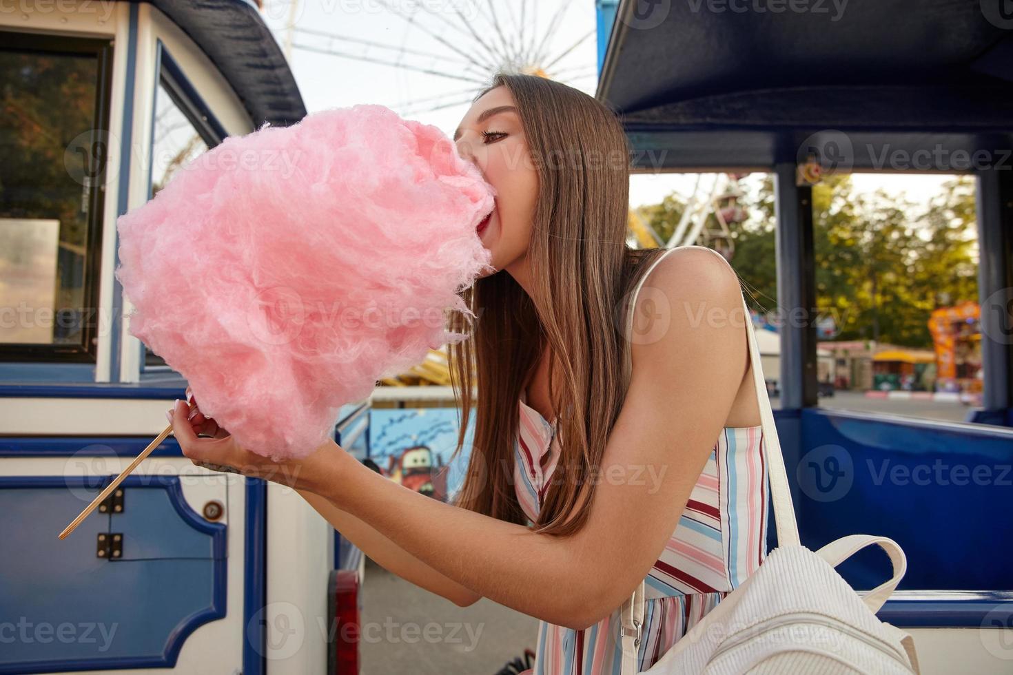 joven y atractiva mujer de cabello largo con cabello castaño comiendo algodón de azúcar rosa en un palo mientras camina por el parque de diversiones, usando un vestido de verano con correas y una mochila blanca foto
