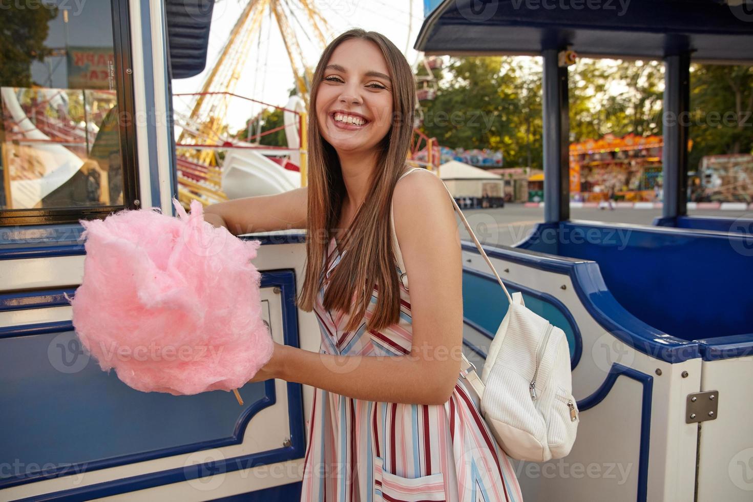 Indoor shot of happy young long haired pretty woman in light summer dress standing over amusement park on warm day with cotton candy in hand, looking at camera happily and smiling widely photo