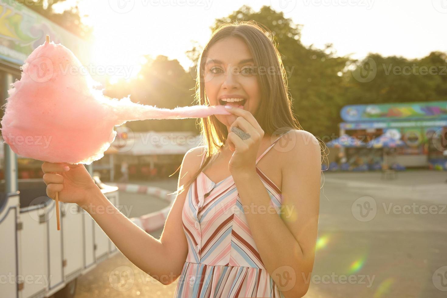 jovencita morena positiva y atractiva con vestido ligero de verano parada sobre un parque de diversiones en un día cálido y soleado, mirando a la cámara alegremente y tirando algodón rosa con la mano en la boca foto