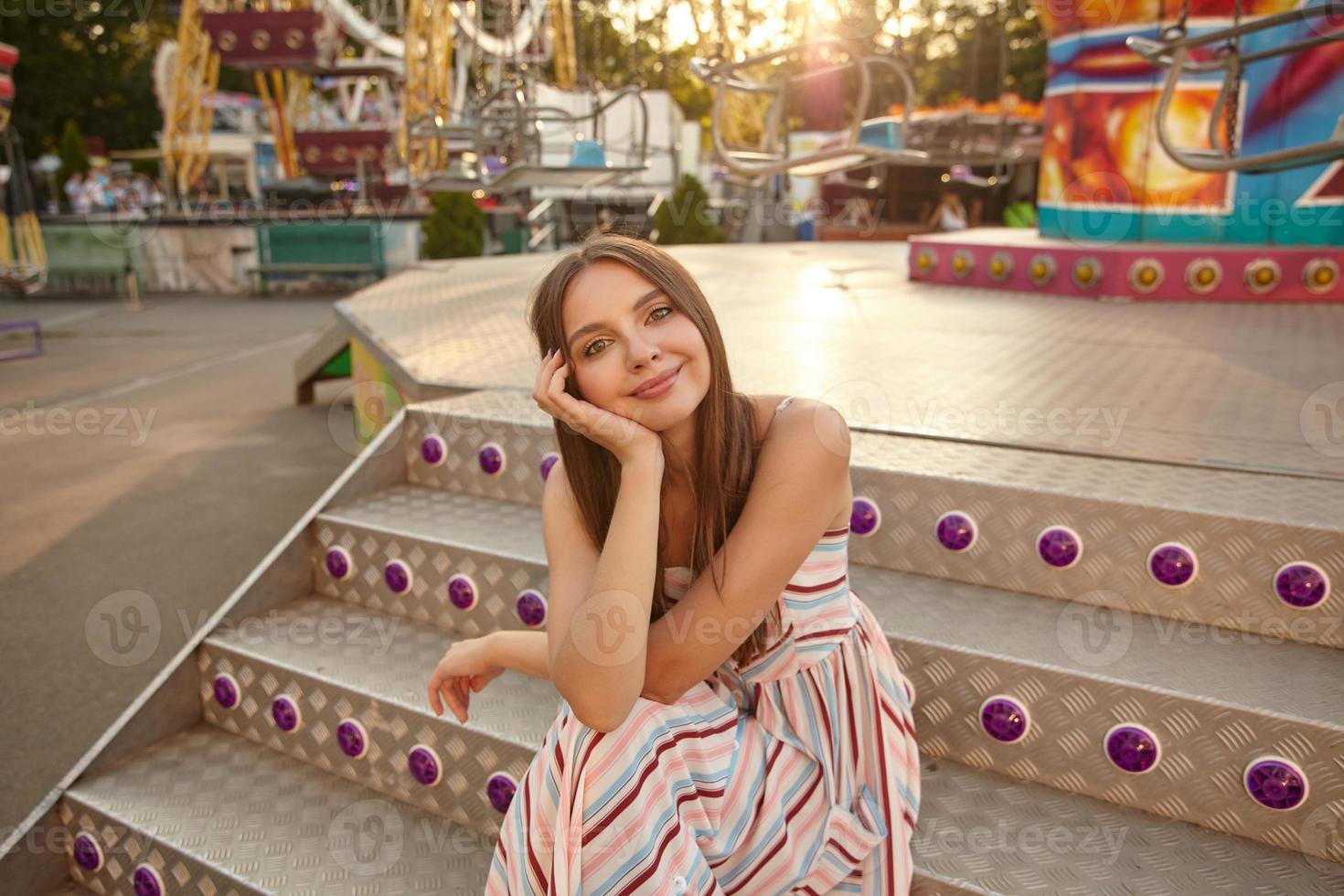 Positive beautiful young woman with brown long hair wearing romantic light dress, sitting on stairs over amusement park, leaning head on her palm and looking to camera with soft smile photo