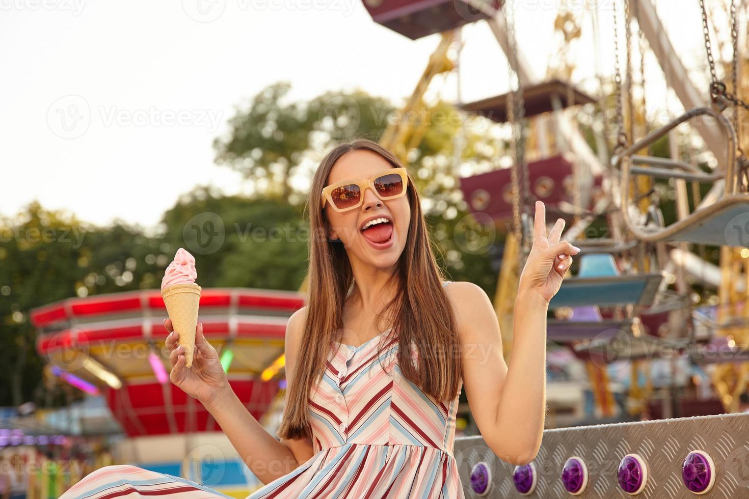 Positive young long haired woman in sunglasses looking to camera with wide mouth opened, holding ice cream cone in hand and raising two fingers in victory sign photo