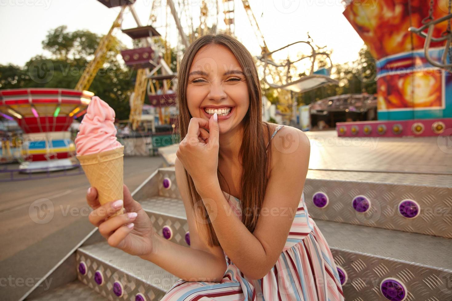 Cheerful young beautiful female with long brown hair sitting over park of attractions with ice cream in hand, looking to camera with broad smile and keeping thumb on her underlip photo