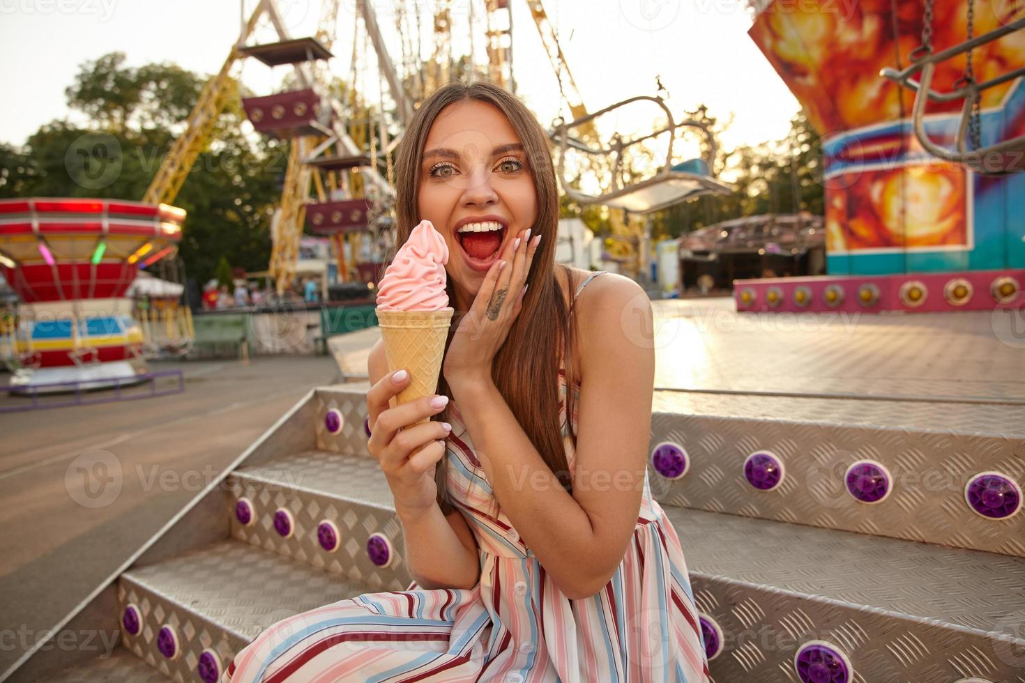 Outdoor shot of joyful attractive brunette female eating ice cream in park of attractions, holding palm on her face and laughing with wide mouth opened, wearing light summer dress photo