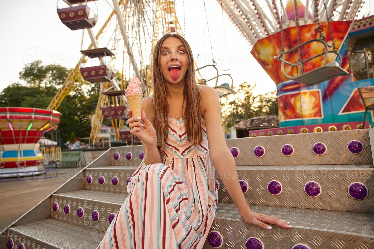 Funny outdoor photo of lovely young female in summer dress posing over ferris wheel in amusement park, holding ice cream in hand and showing pink tongue