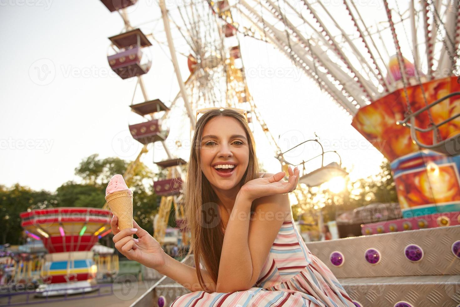 feliz hermosa mujer joven con el pelo largo y castaño posando sobre la rueda de la fortuna en un cálido día de verano, manteniendo el cono de helado en la mano y levantando la palma, mirando la cámara con alegría foto