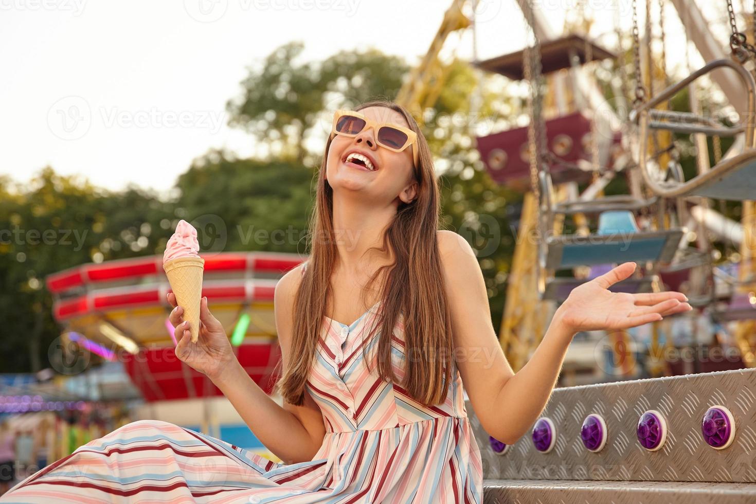 Happy young beautiful woman in romantic light dress sitting over amusement park decorations with ice cream cone in hand, smiling with closed eyes and raising palm up photo