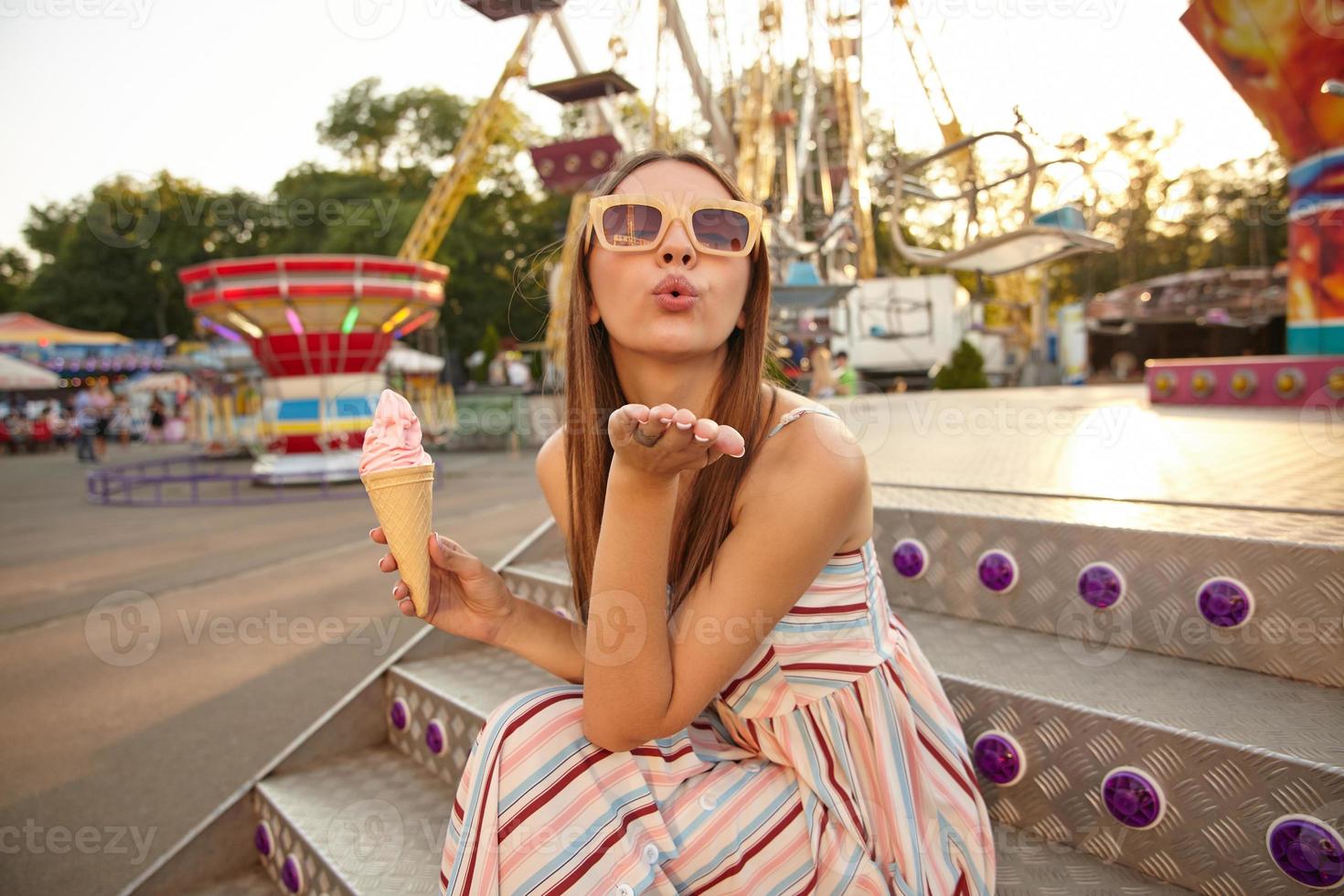 Portrait of atrractive young brunette woman with long hair posing outdoor on warm sunny day, sitting on stairs and holding ice cream in cone, raising palm and blowing air kiss to camera photo