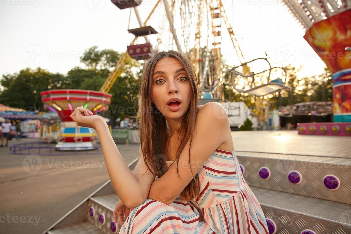 Outdoor shot of pretty young woman with brown hair in summer light dress looking to camera with surprised face, sitting on stairs with hands on her knees, contracting forehead with wide mouth opened photo