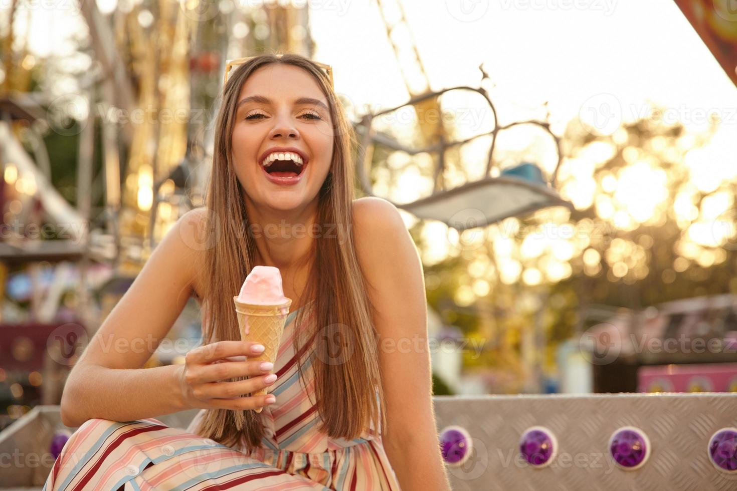 Portrait of joyful young lovely woman with sunglasses on her head posing outdoor over park of attractions, laughing with wide mouth opened and keeping ice cream cone in hand photo