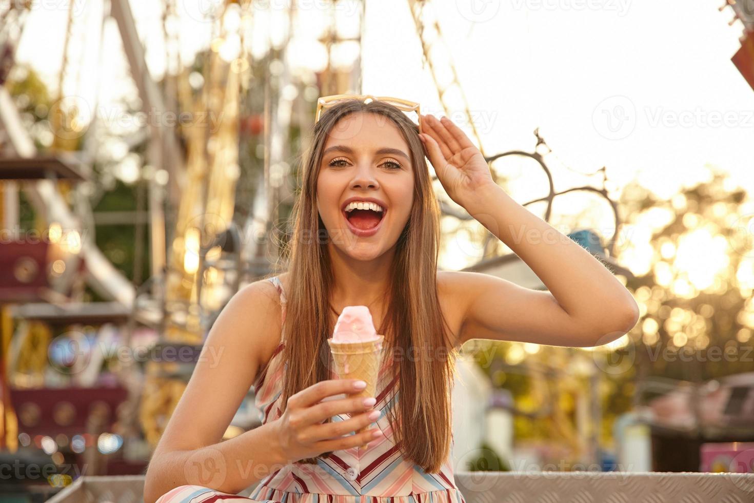 Young cheerful long haired pretty lady with sunglasses on her head looking at camera with broad smile, eating ice cream cone while walking through park of attractions, wearing summer clothes photo
