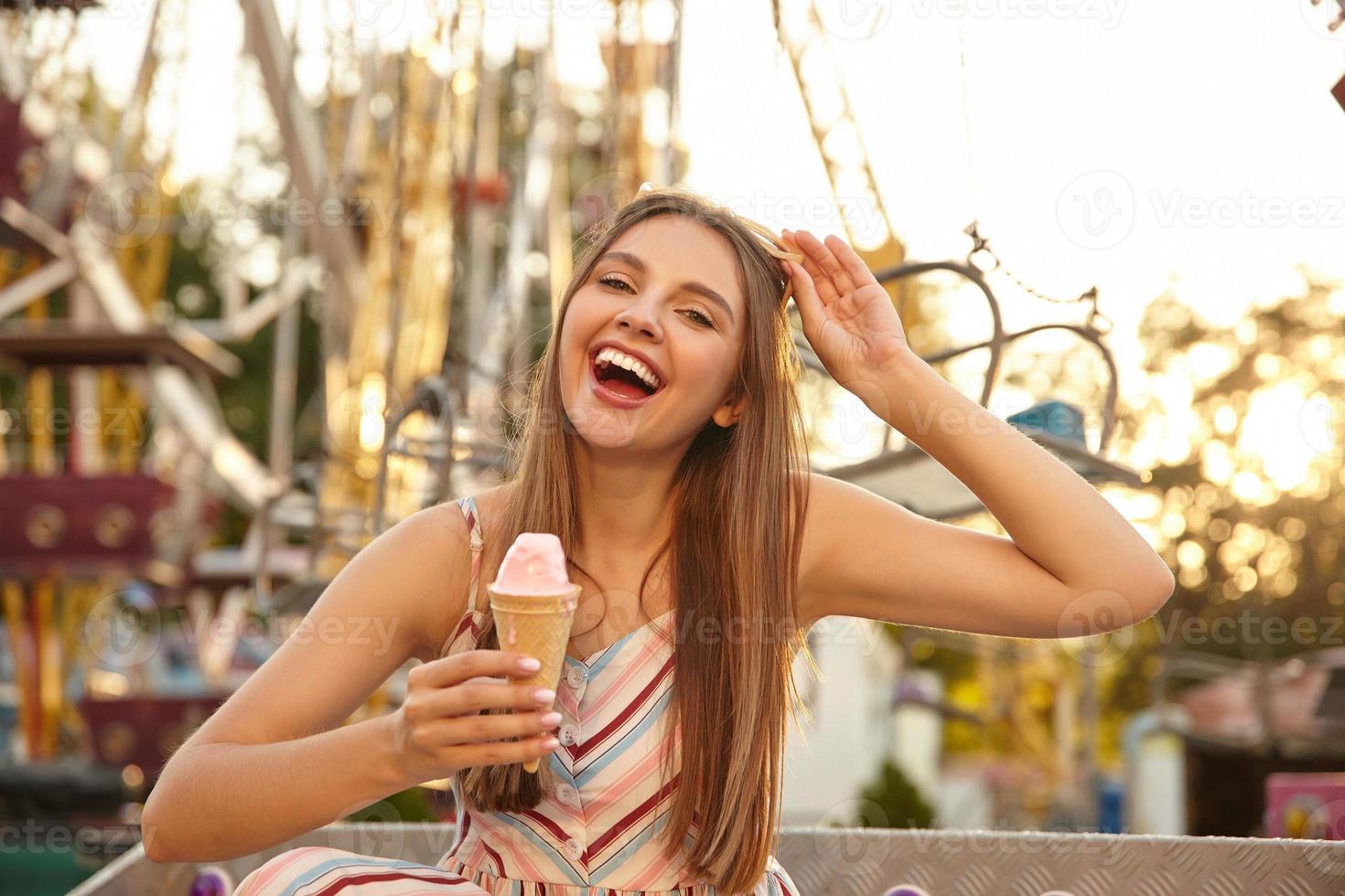 Outdoor shot of pretty young happy female with long hair eating ice cream in cone while walking through amusement park, laughing joyfully with wide mouth opened and taking sunglasses off photo