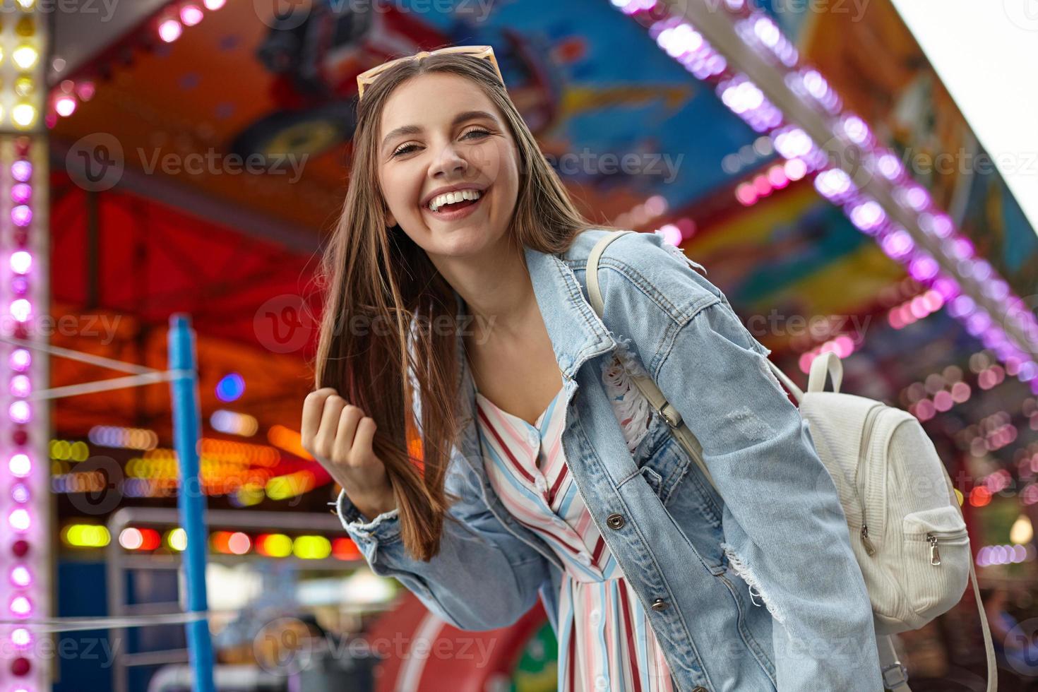 Cheerful young pretty woman with long brown hair walking in park of attractions, wearing casual clothes and white backpack, holding her hair and looking to camera with wide sincere smile photo