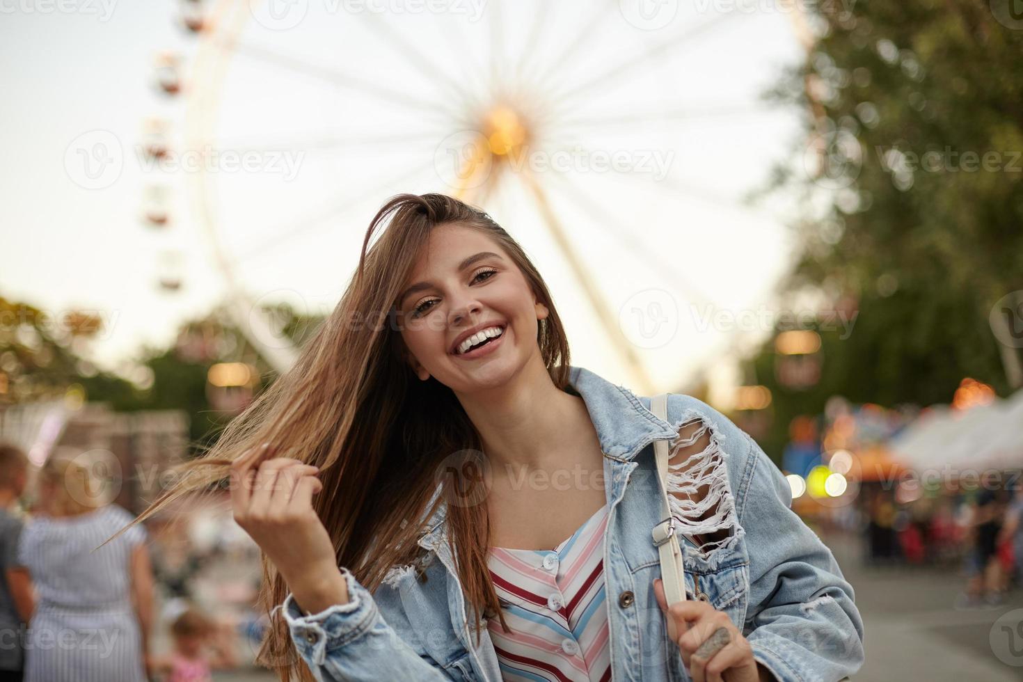 Charming pretty young lady with long brown hair posing over ferris wheel in casual clothes, waving her hair and smiling cheerfully to camera, positive emotions concept photo