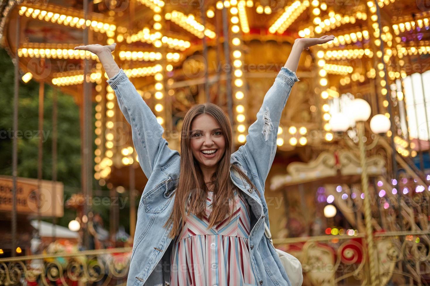 Outdoor photo of pretty young female with long brown hair looking to camera with sincere smile and raising hands happily, wearing romantic dress and jeans coat