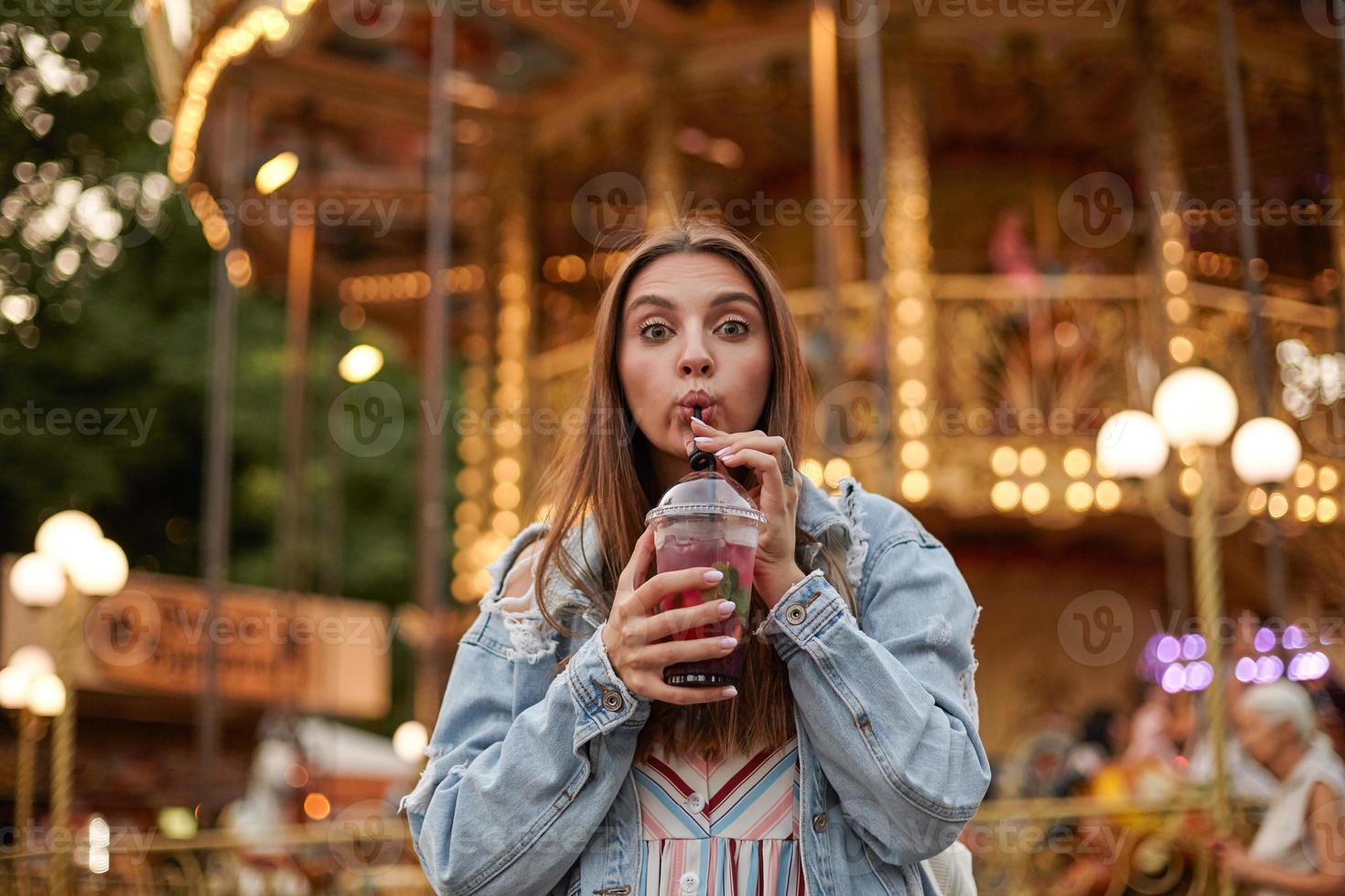 Outdoor photo of lovely long haired brunette woman in casual clothes drinking lemonade with straw while walking in park of attractions, looking at camera and contracting forehead