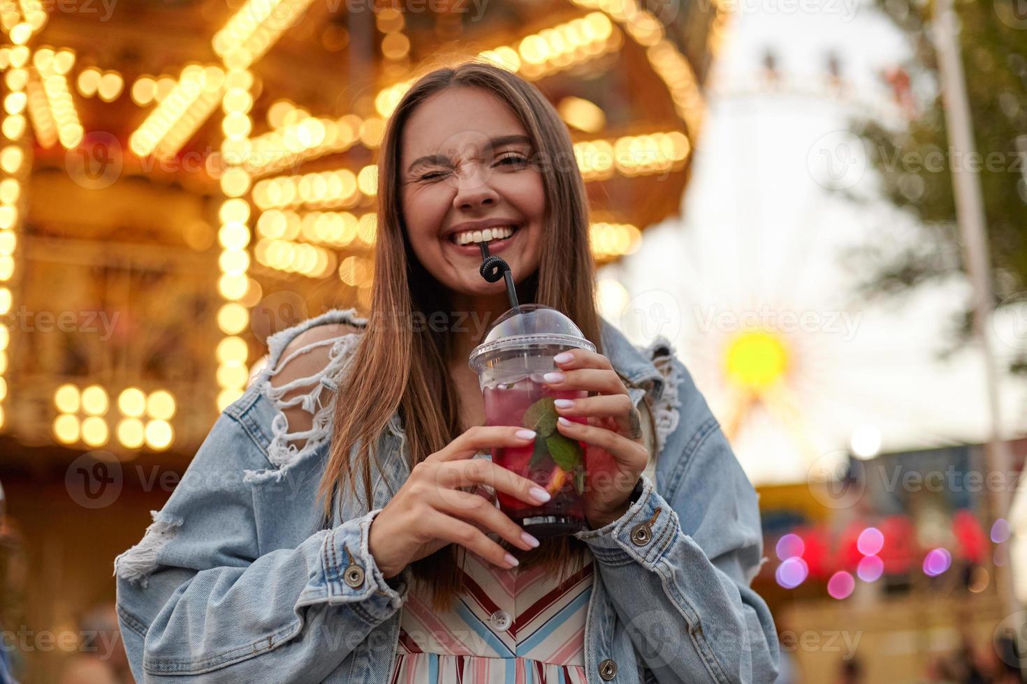 Positive long haired young pretty female in trendy jeans coat posing over park of attractions, giving wink and smiling widely to camera while drinking lemonade with straw photo