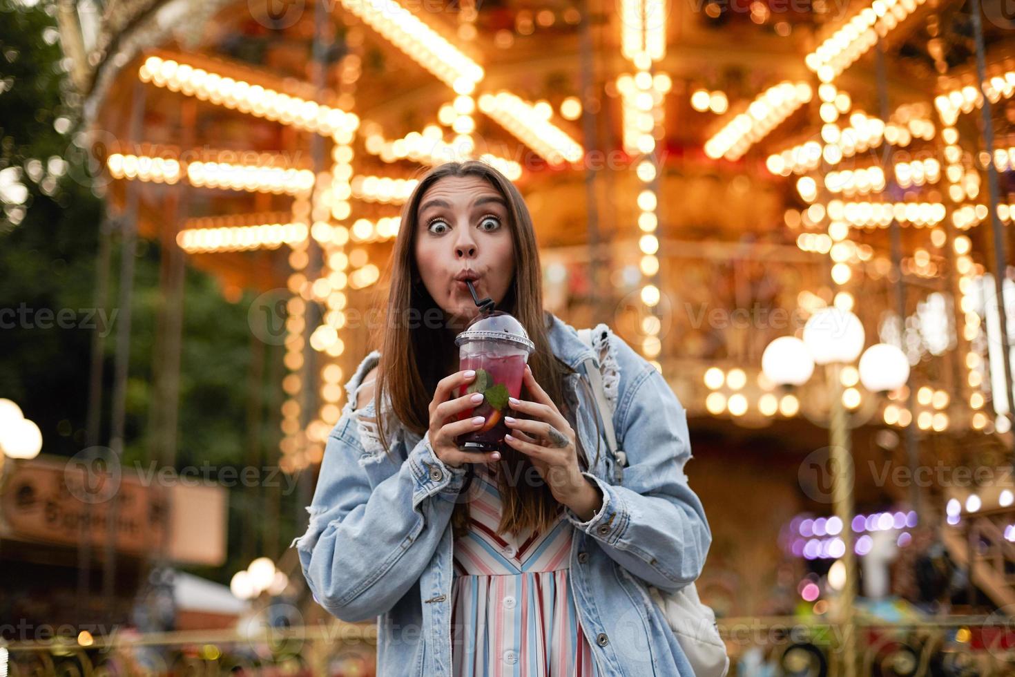 Outdoor photo of lovely young female with long brown hair looking to camera with eyes wide open and contracting forehead, drinking lemonade while walking through park of attractions