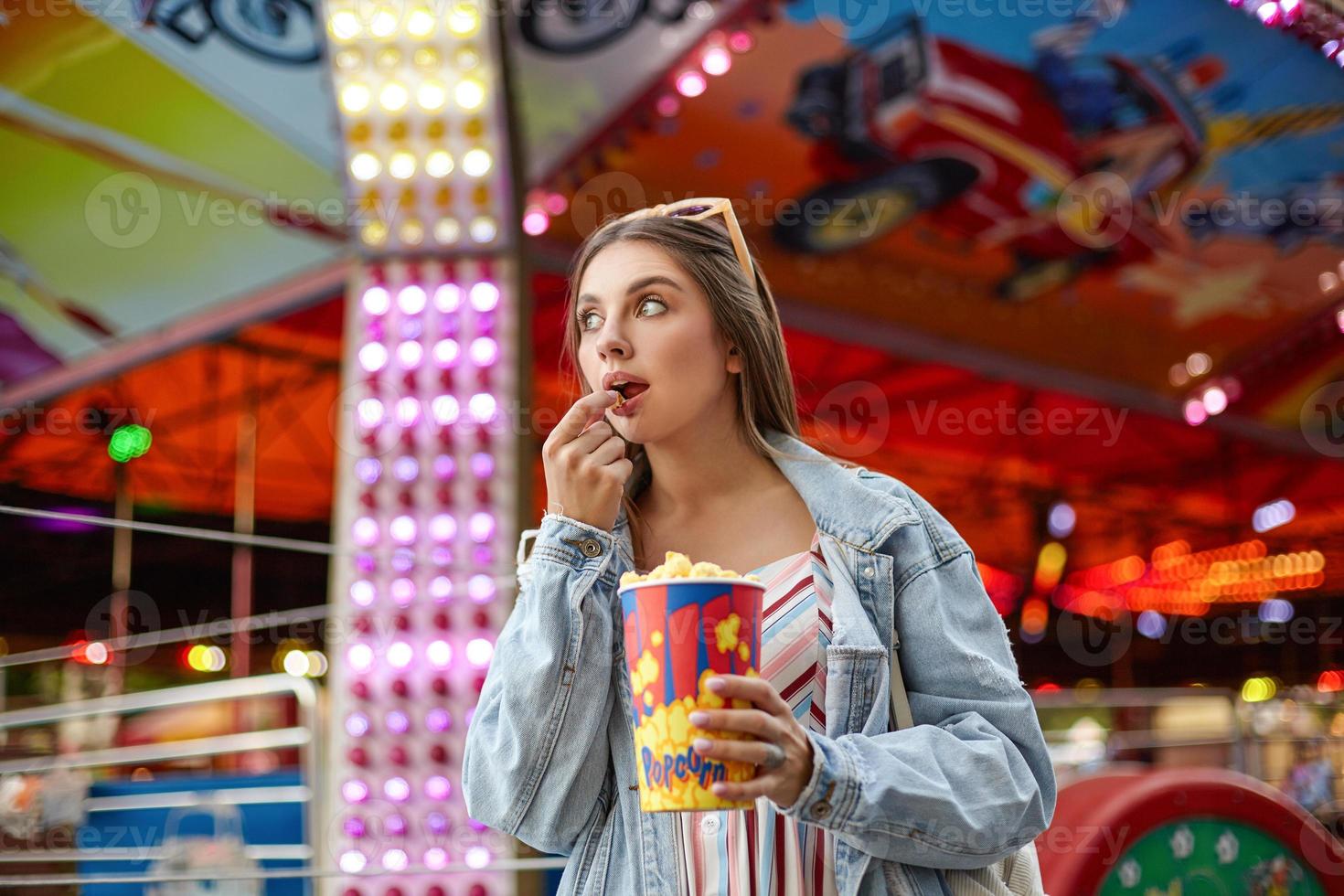 Portrait of attractive young brunette woman with sunglasses on her head eating popcorn while walking in amusement park, looking aside with interest photo