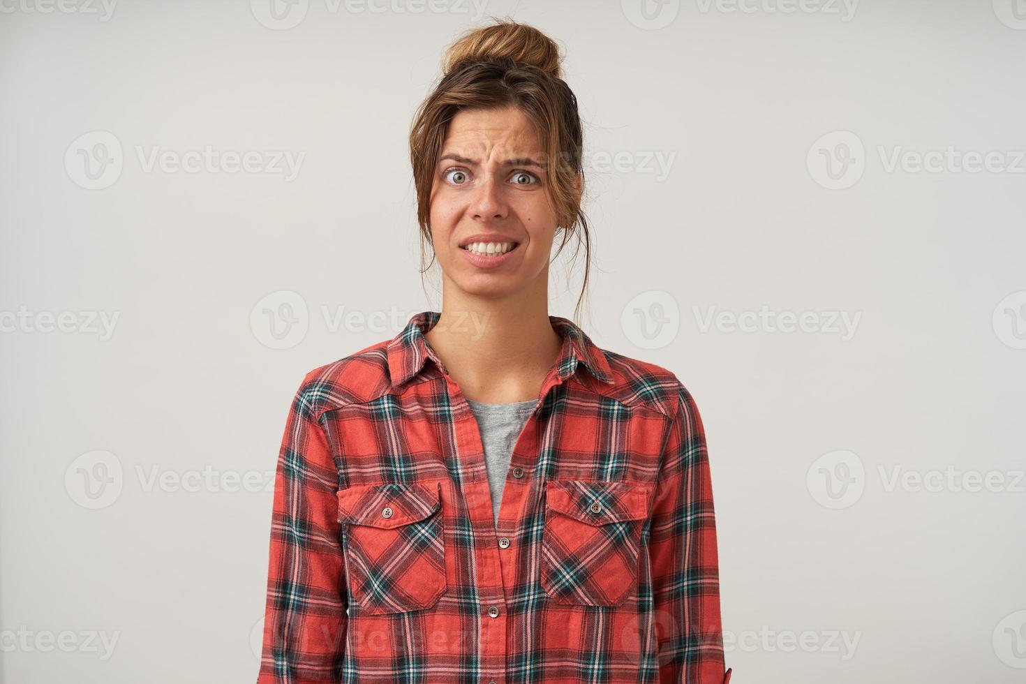 Studio photo of dark haired lovely woman standing over white background with confused face, wearing casual clothes, showing teeth and wrinkling forehead