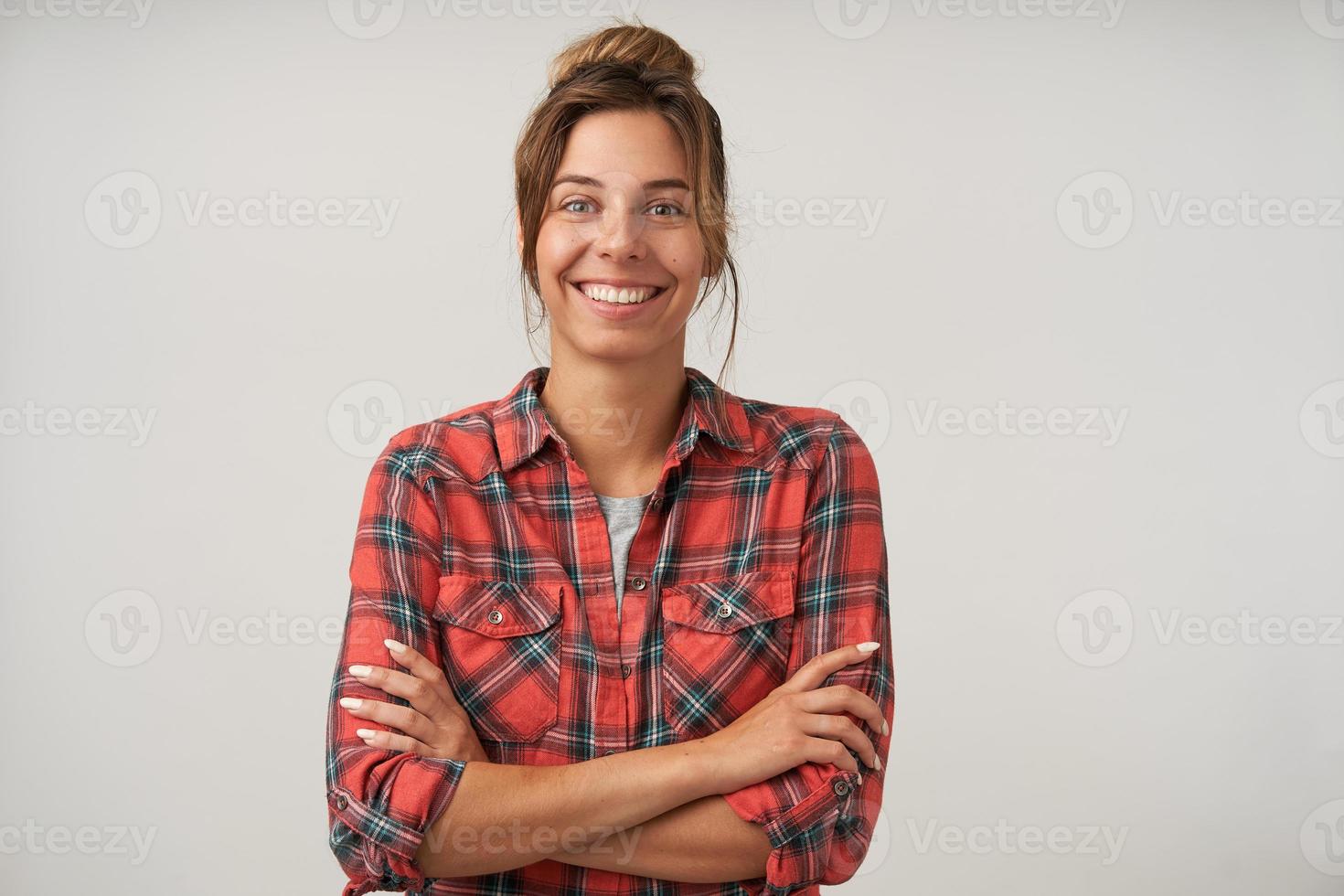 Studio portrait of young pretty dark haired female with bun hairstyle wearing casual clothes, posing over white background, looking happily to camera with crossed arms on chest photo