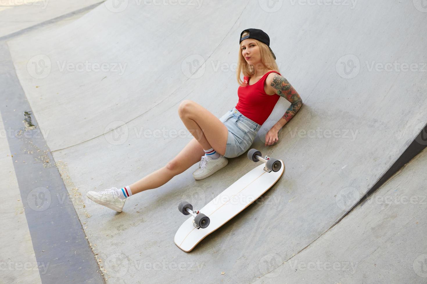 Attractive young blonde woman with tattoos in jeans shorts and red top sitting over stunt park, grimacing on camera while taking rest after riding skate board photo