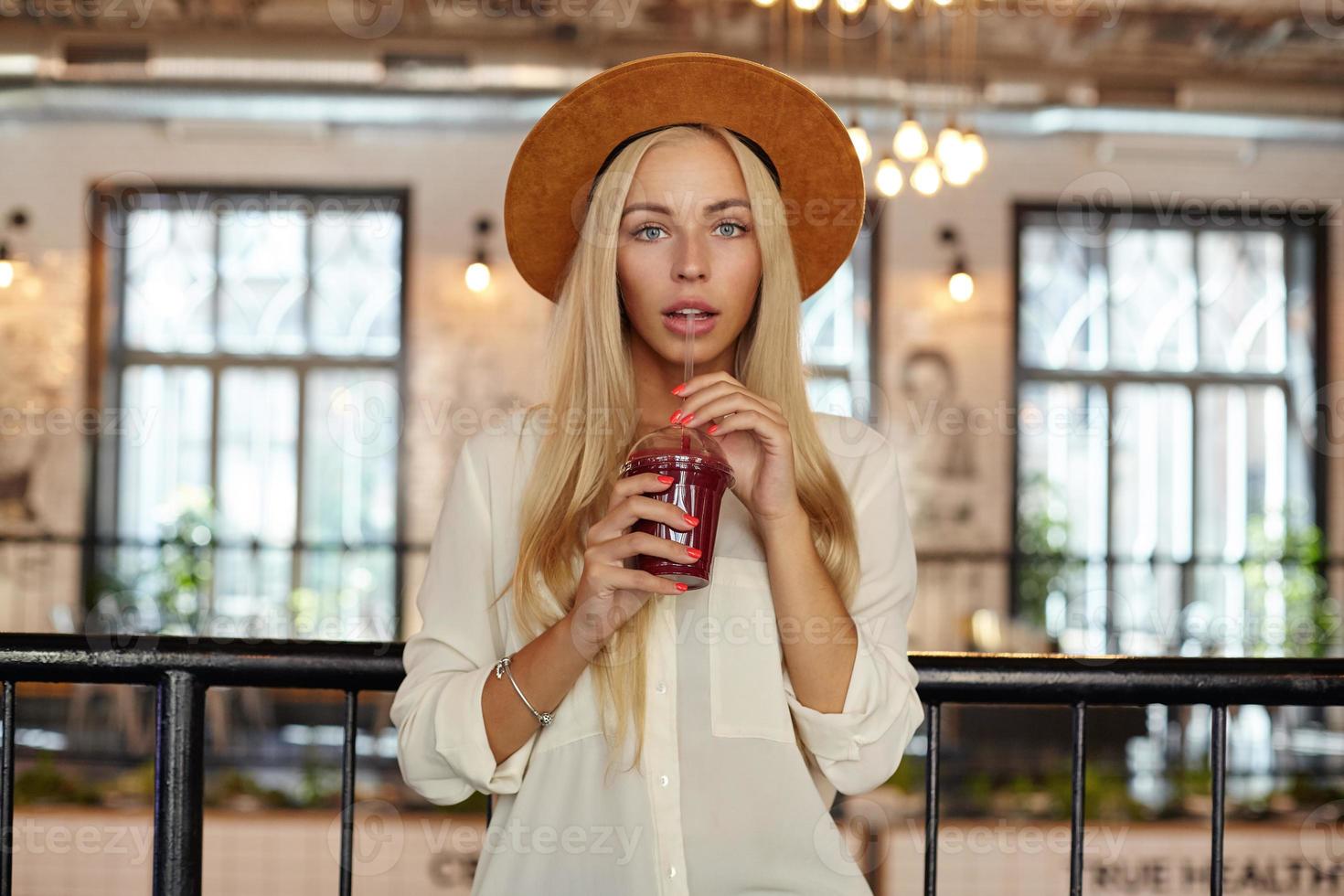 toma interior de una hermosa mujer rubia de cabello largo con ojos azules posando sobre el interior del restaurante, bebiendo limonada con paja, usando sombrero y camisa foto