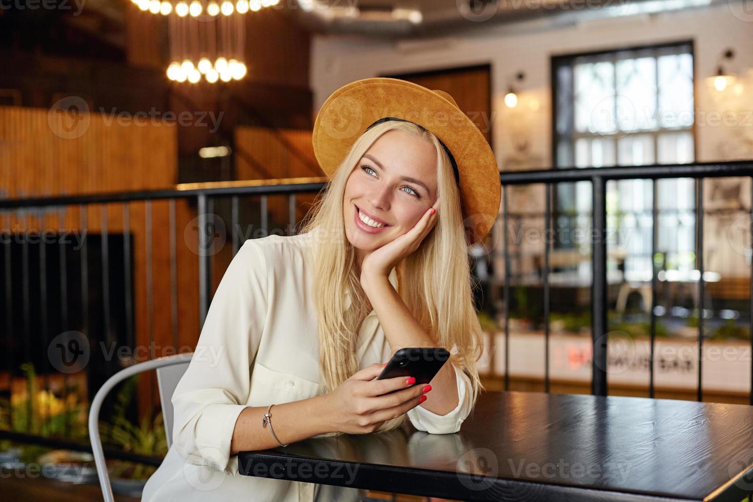 foto interior de una encantadora joven rubia con pantalones blanco y sombrero marrón sentada en el interior de un café, inclinando la cabeza sobre su palma y mirando soñadoramente a un lado