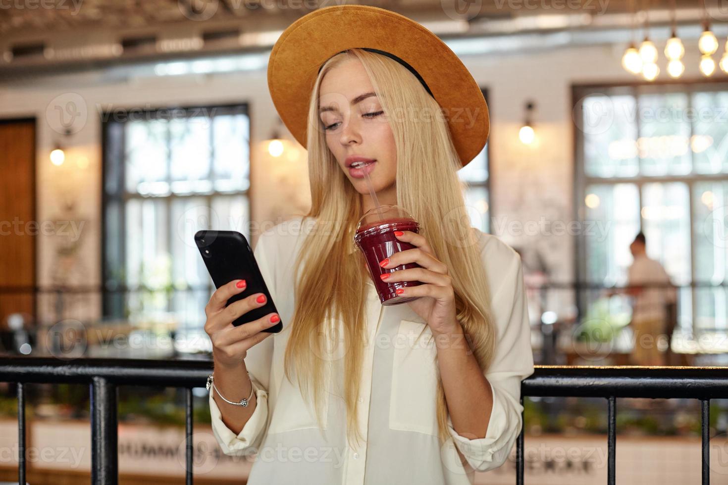 Young pretty blonde female in white shirt and wide hat drinking lemonade while lunch break, holding mobile phone in hand and looking at screen with calm face photo