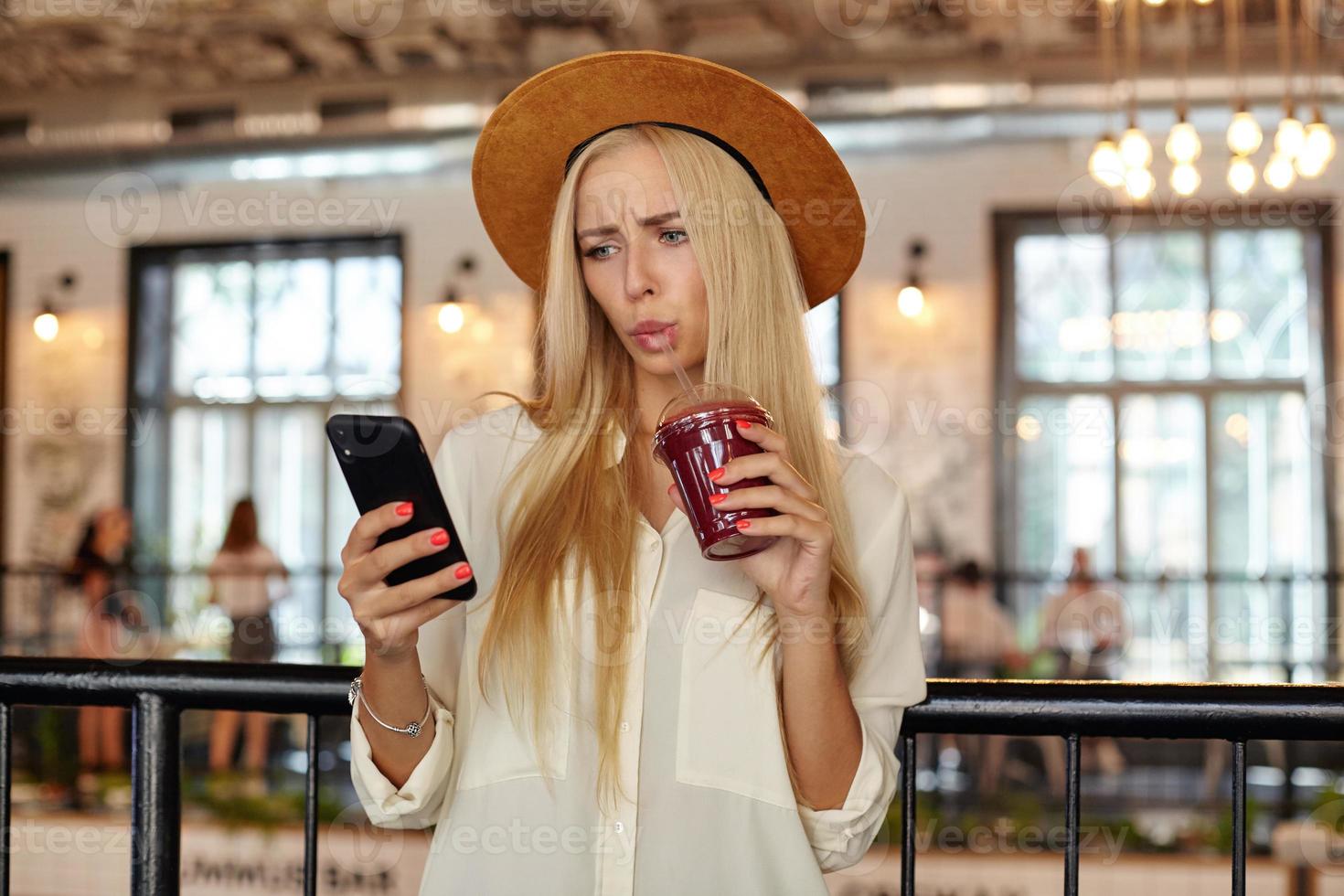 Indoor photo of beautiful confused woman with long hair posing over restaurant interior with cup of lemonade in hand, looking at her mobile phone and reading messages