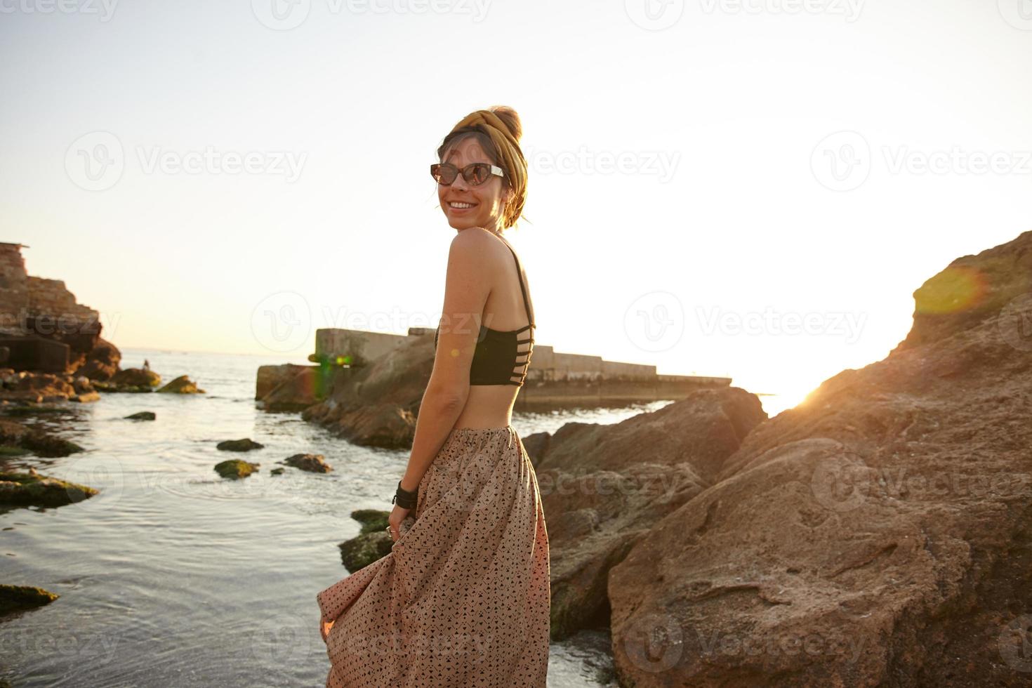 foto al aire libre de una feliz mujer de cabello oscuro parada junto al mar temprano en la mañana, usando ropa informal y gafas de sol, mirando a la cámara y sonriendo alegremente