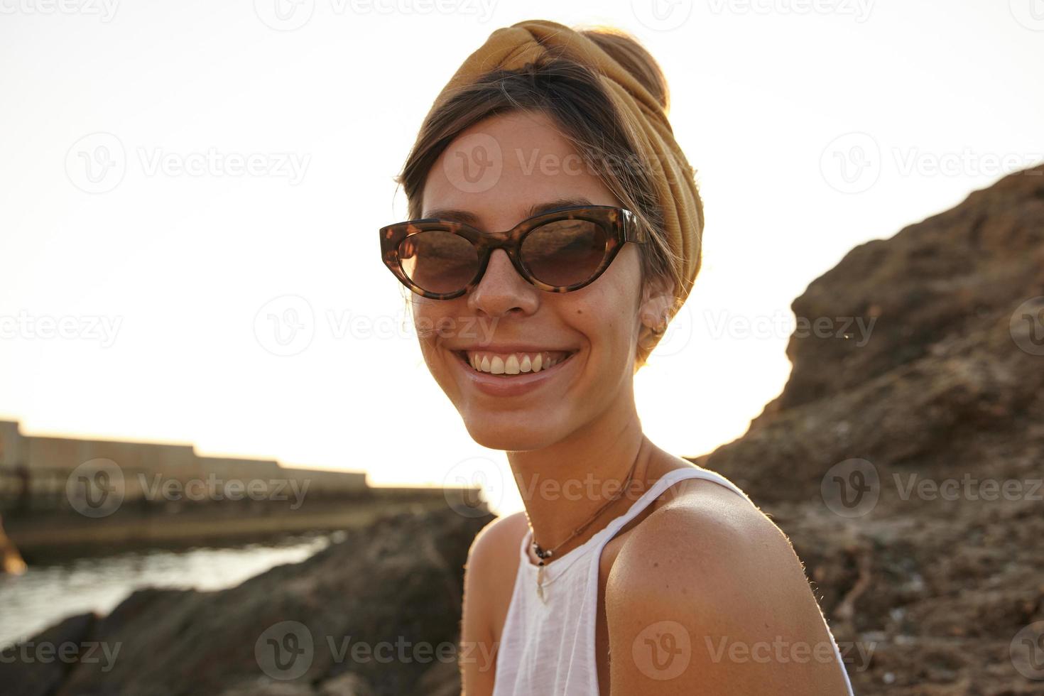 Outdoor close up of beautiful dark haired woman in headband and sunglasses looking to camera with charming smile, posing over seaside view on warm summer day photo