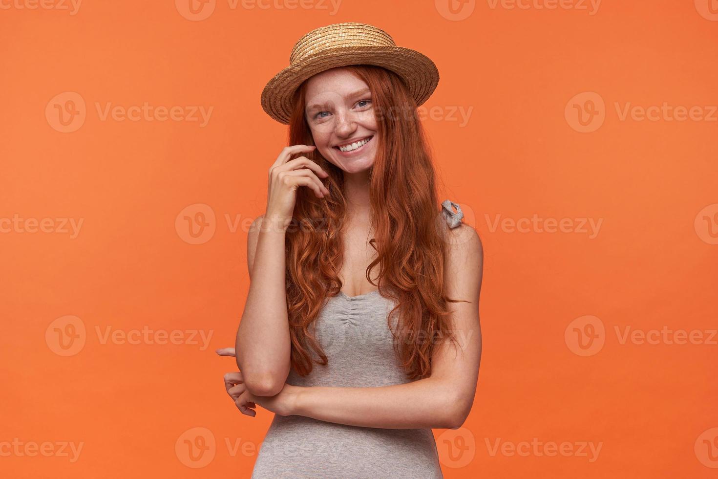 foto de estudio de una joven y bonita mujer de pelo rojo con ropa informal y sombrero de navegante, parada sobre un fondo naranja, mirando a la cámara con una sonrisa encantadora y tocando su cabello ondulado