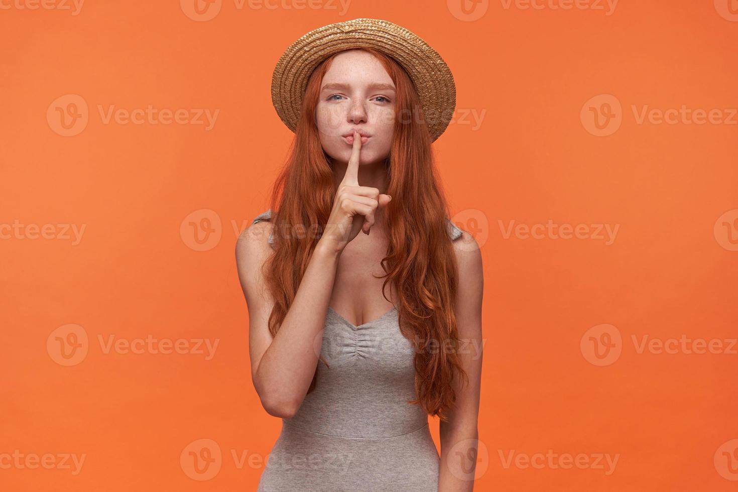 Portrait of charming young woman with wavy foxy hair raising forefinger to her lips, asking to keep silence, posing over orange background in grey shirt and straw hat photo