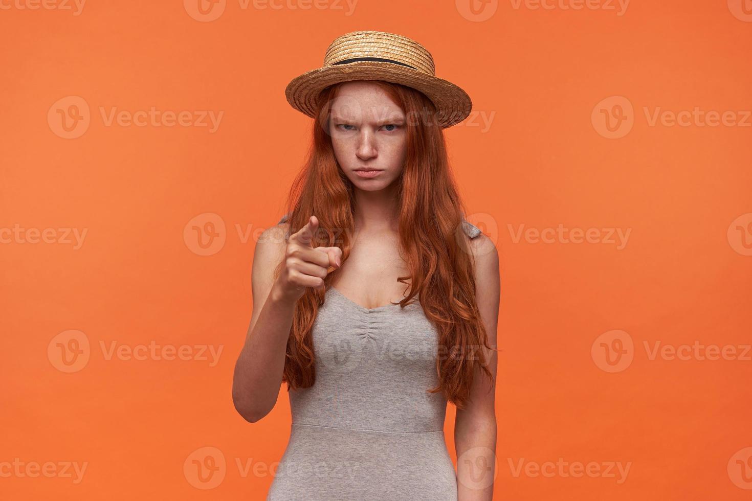 retrato de una joven pelirroja frunciendo el ceño con el pelo largo con camisa gris y sombrero de paja, levantando el dedo índice en señal de advertencia, posando sobre fondo naranja foto