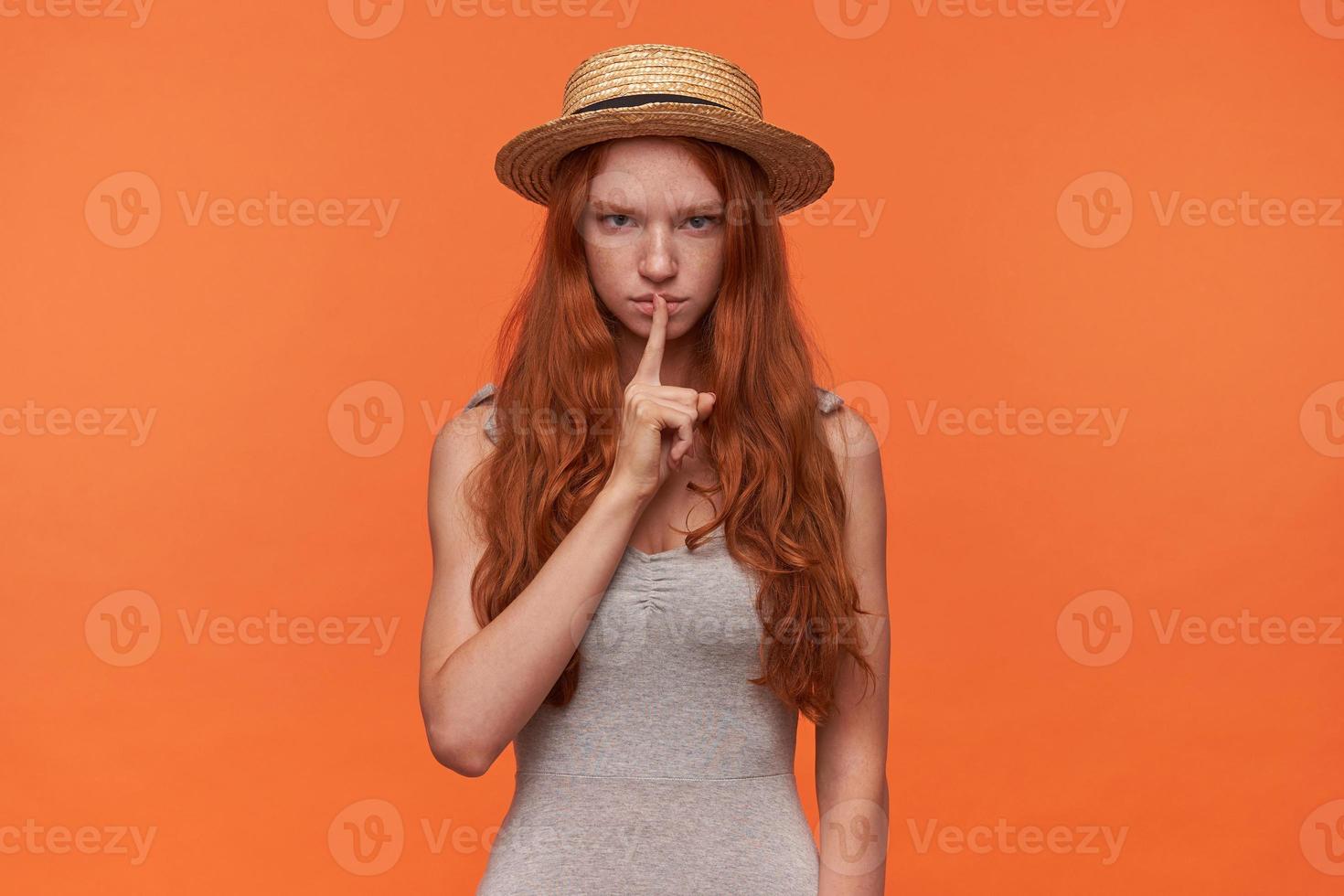 Studio photo of beautiful young foxy lond haired female raising index finger in hush gesture, looking to camera with serious face and calling for silence, isolated over orange background