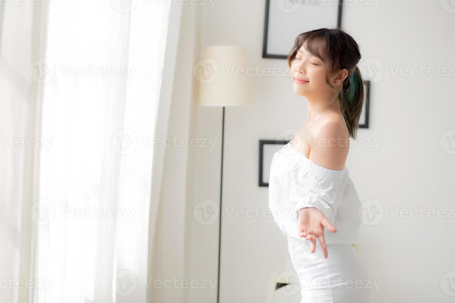 Portrait of beautiful young asian woman standing the window and smile while wake up with sunrise at morning, girl happy with freshness and cheerful, lifestyle and relax concept. photo