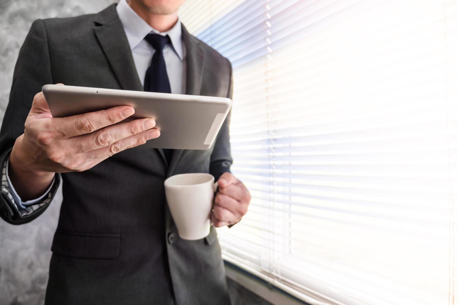 Close up of businessman using tablet and holding coffee cup while standing at a window in an office photo