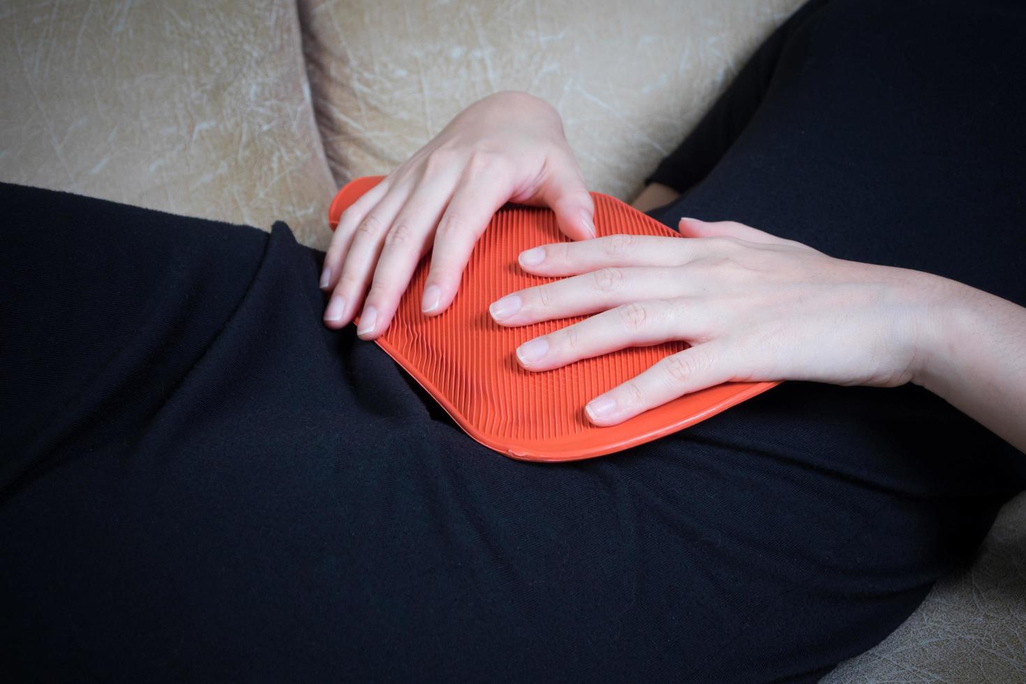 Sick woman lying on sofa with hot water bag and holding it on her belly photo