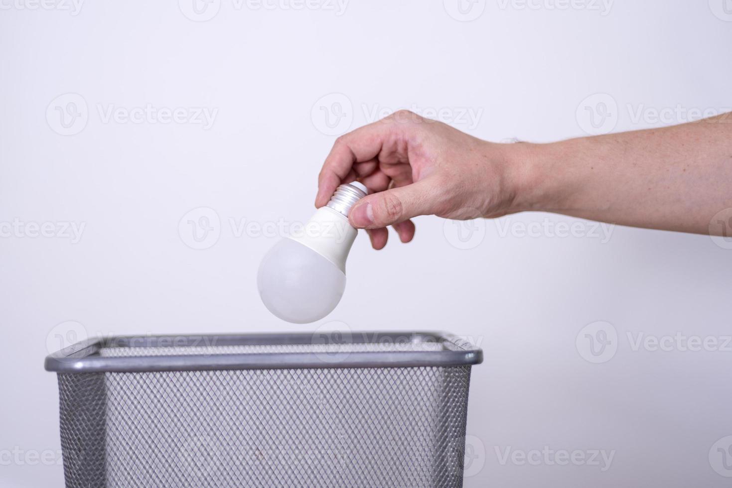 A hand throws a light bulb into a trash can against a gray background