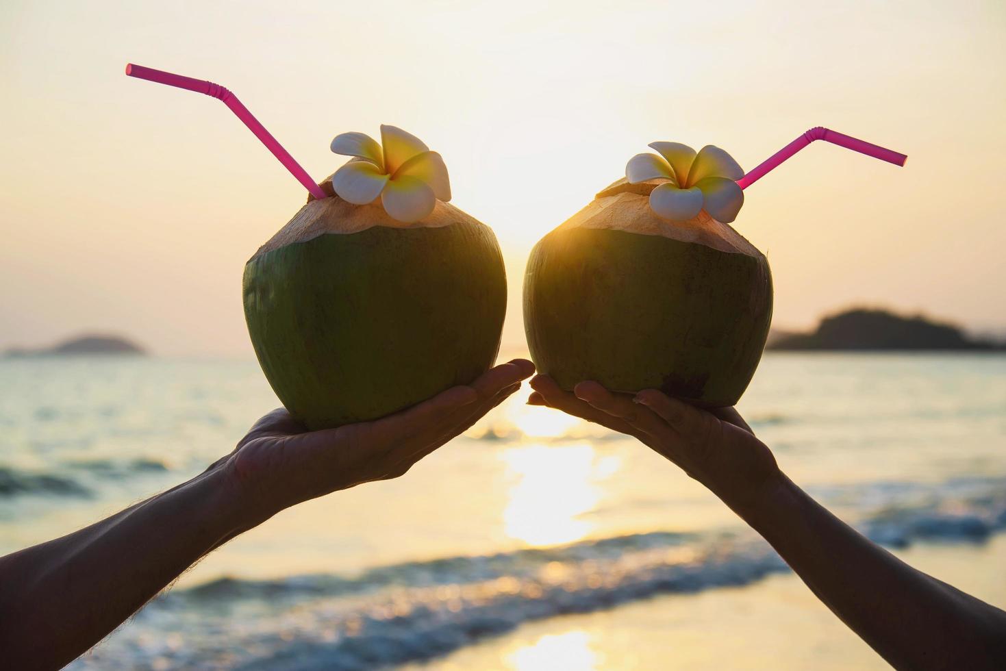 Silhouette of fresh coconut in couples hands with plumeria decorated on beach with sea wave background - tourist with fresh fruit and sea sand sun vacation background concept photo